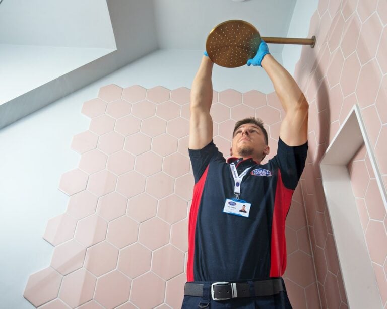 A plumber stretching to check the top of a gold shower head on a pink tiled wall