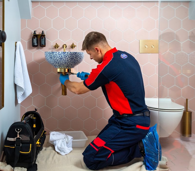 A plumber using a wrench underneath a bathroom sink with a tool bag next to him