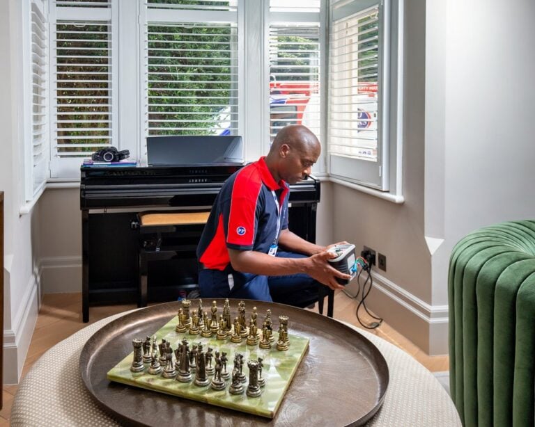 An electrician in a living room, in front of a grand piano, testing the socket with a handheld device