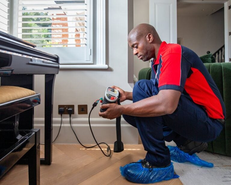 An electrician with shoe covers on inspects a handheld device linked to a living room plug socket
