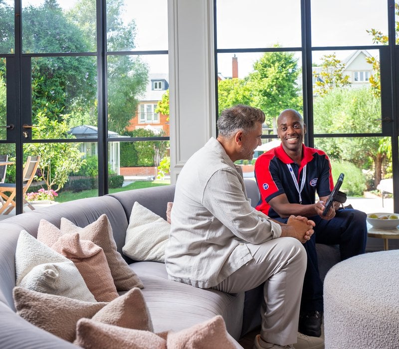 A smiling engineer in Pimlico Plumbers uniform, sat with a homeowner on a sofa